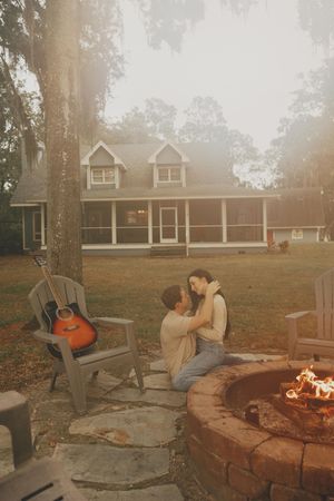 A cozy outdoor scene with a stone fire pit and Adirondack chairs at sunset with a rustic house in the background.