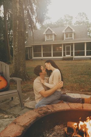 Couple relaxing and cuddling near a cozy fire pit outside their home at dusk.