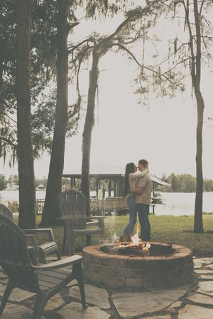 Couple relaxing by a lakeside fire pit with Adirondack chairs and trees at dusk.