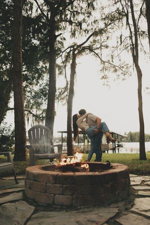 A curved stone fire pit surrounded by Adirondack chairs sits among tall trees at dusk.