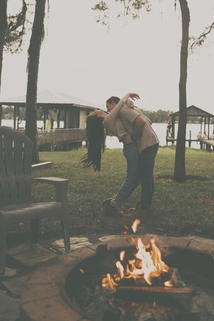 A couple dances playfully near a campfire at dusk with trees in the background.