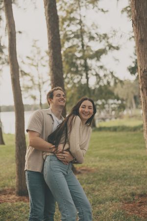 Playful couple laughing together in a grassy area with pine trees in the background.