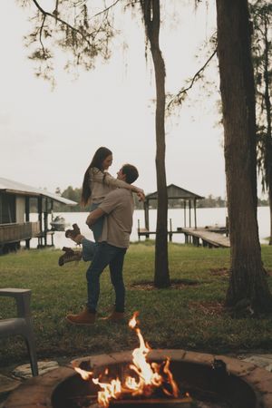 A couple dances and spins together near a lakeside fire pit at sunset.