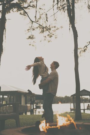 A couple performs a dramatic dance lift near a fire pit with trees in the background.
