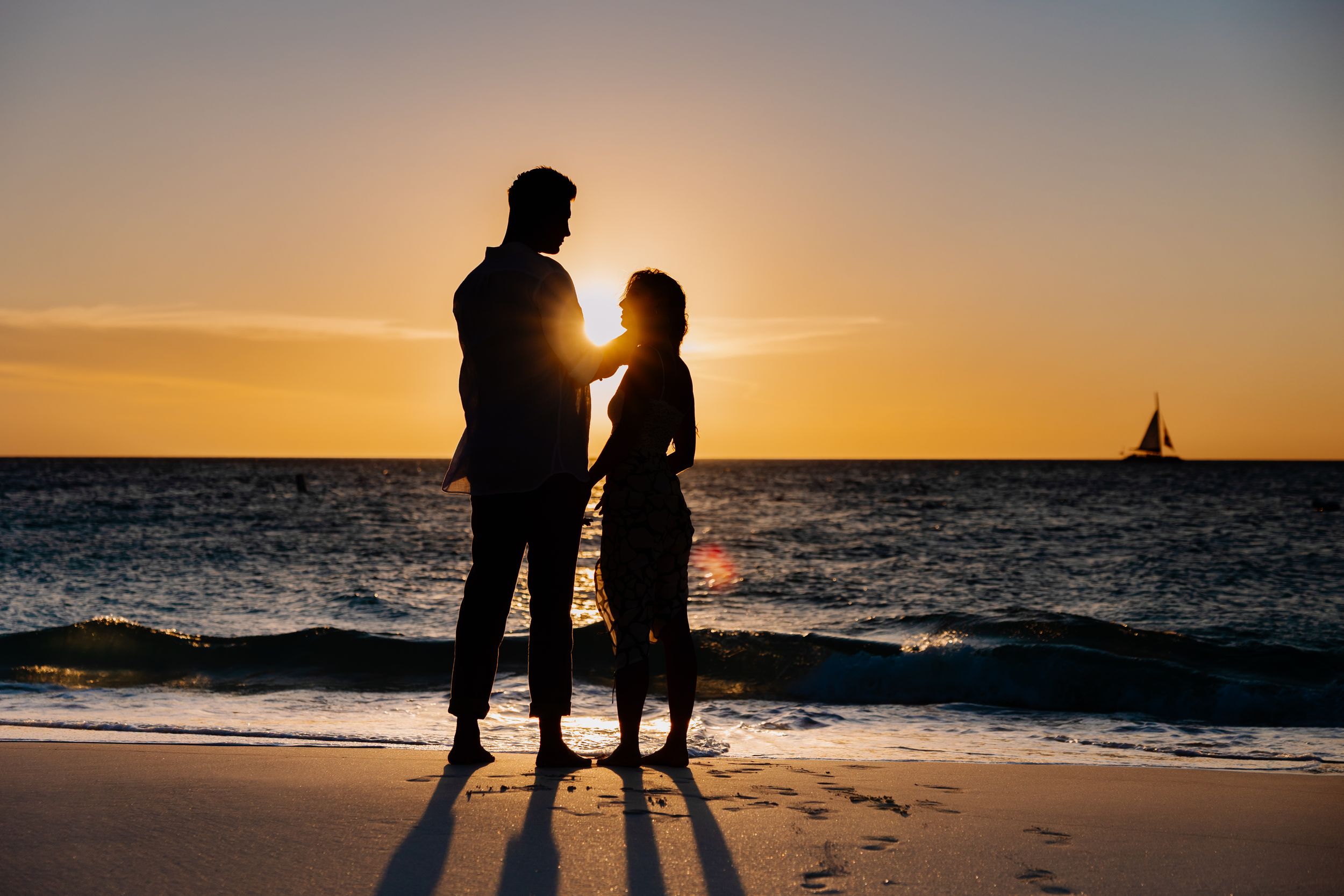 Two silhouettes stand together on a beach at sunset with a sailboat on the horizon.