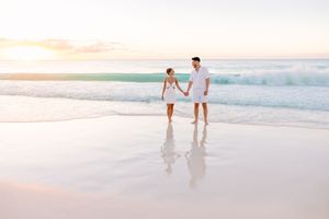 Couple in white outfits walking along a scenic beach at sunset with gentle waves in the background.