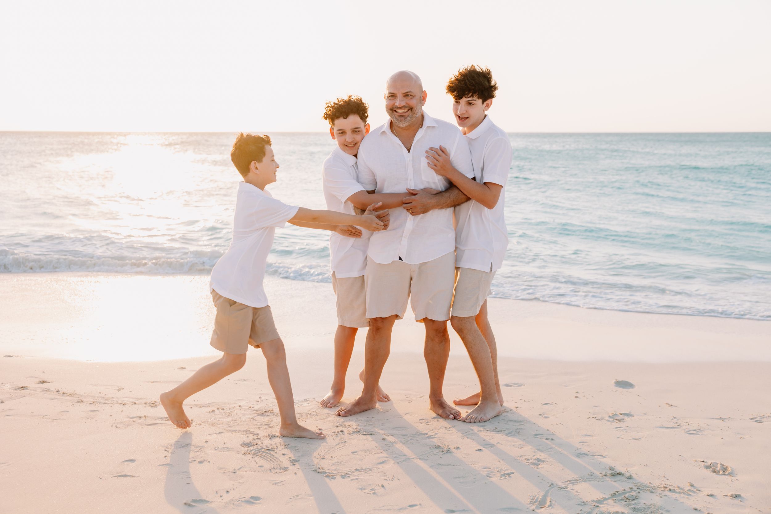 Family of four enjoying a sunny beach day, with boys hugging their grandfather on white sand near calm ocean waves.