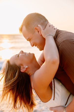 Couple laughing and embracing on a beach at golden sunset, woman leaning back joyfully in mans arms.