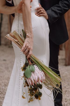 Bride in white gown holding a protea wedding bouquet with tropical greenery while groom embraces her waist.