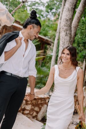 Happy couple in wedding attire holding hands outdoors, bride in white dress and groom in white shirt, tropical setting.