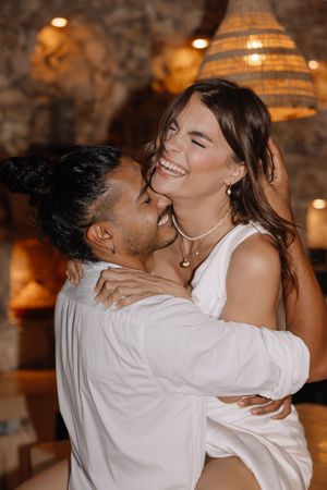 Happy couple embracing and laughing together in a warmly lit rustic venue, both dressed in white outfits.
