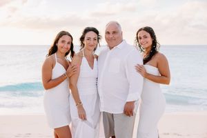 Family of four posing together on a sandy beach in matching white outfits with ocean waves in the background.