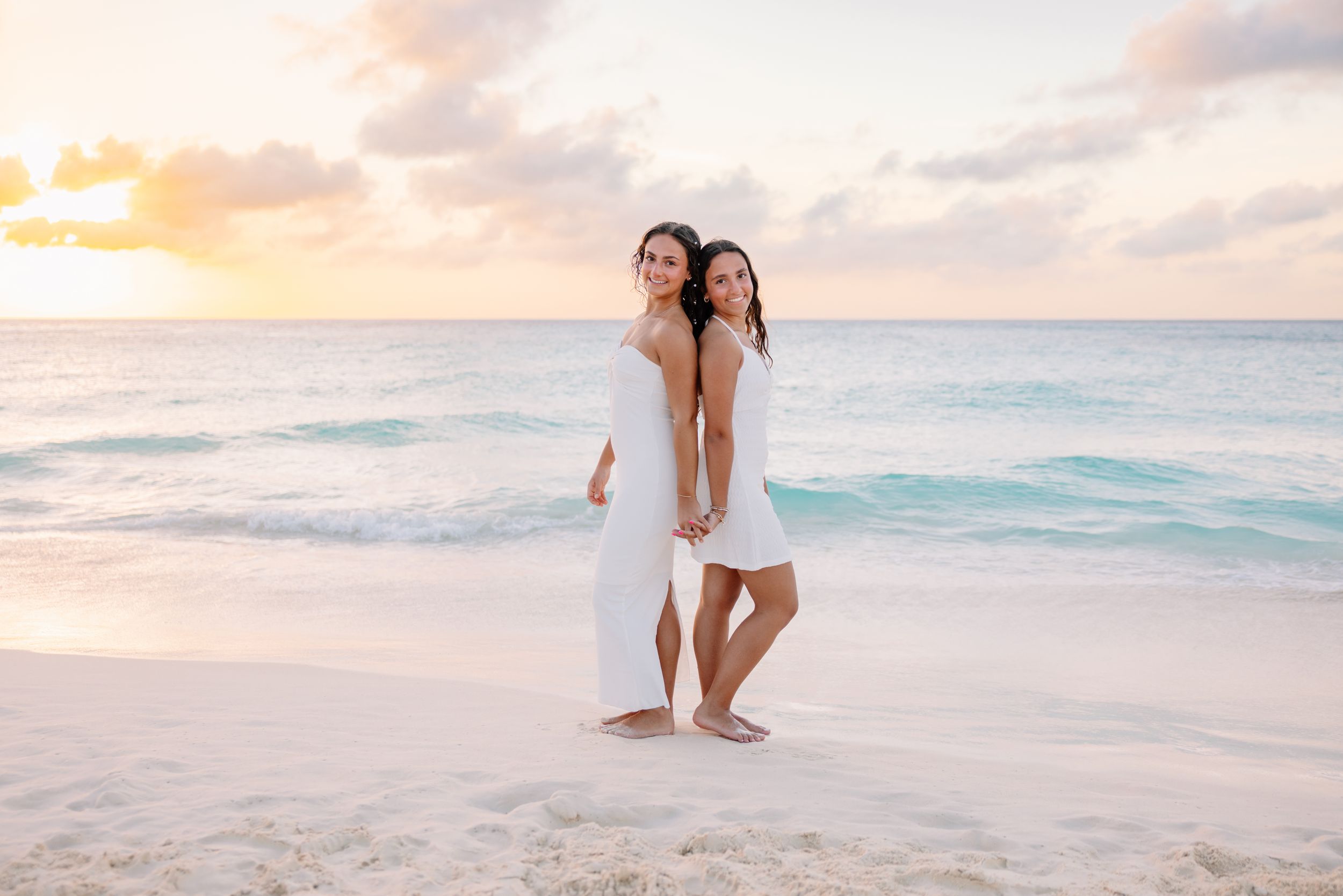 Two women in white dresses hold hands on a tropical beach at sunset with turquoise waves behind them.