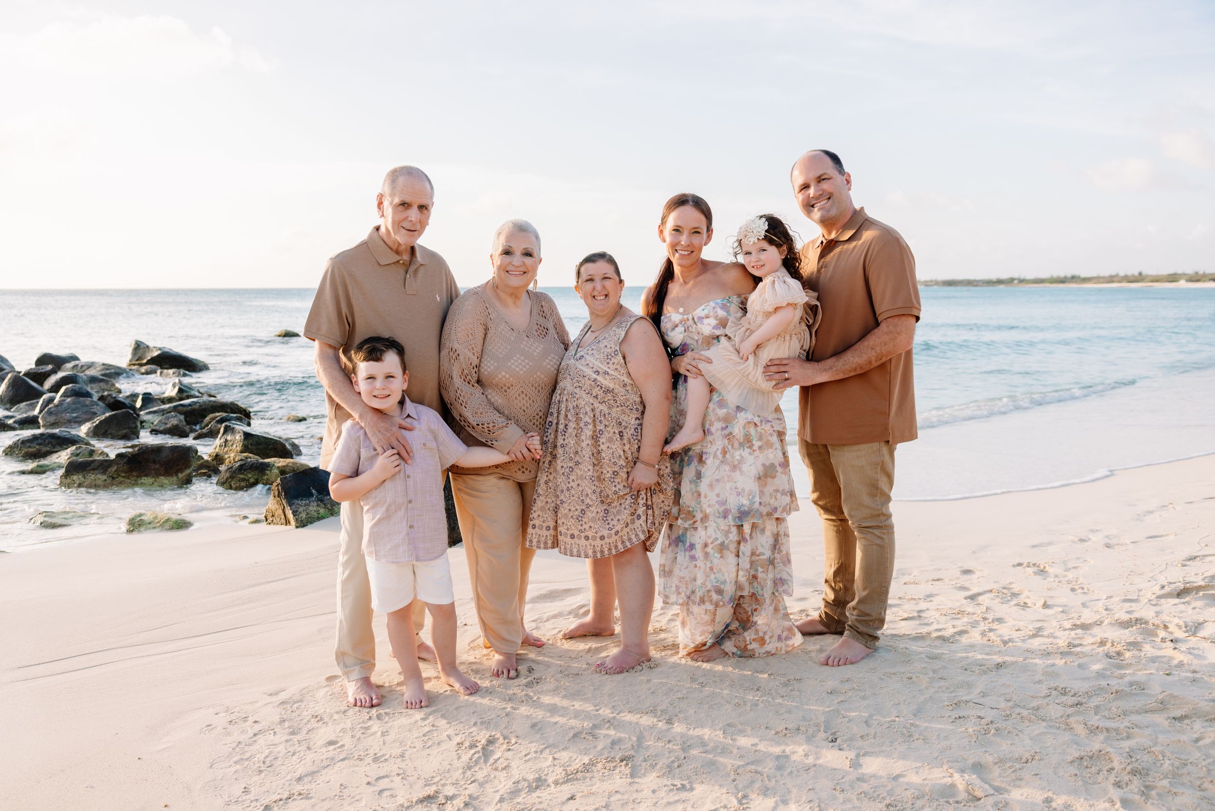 Family of seven posing together on a sandy beach with ocean waves and rocky shore in the background.