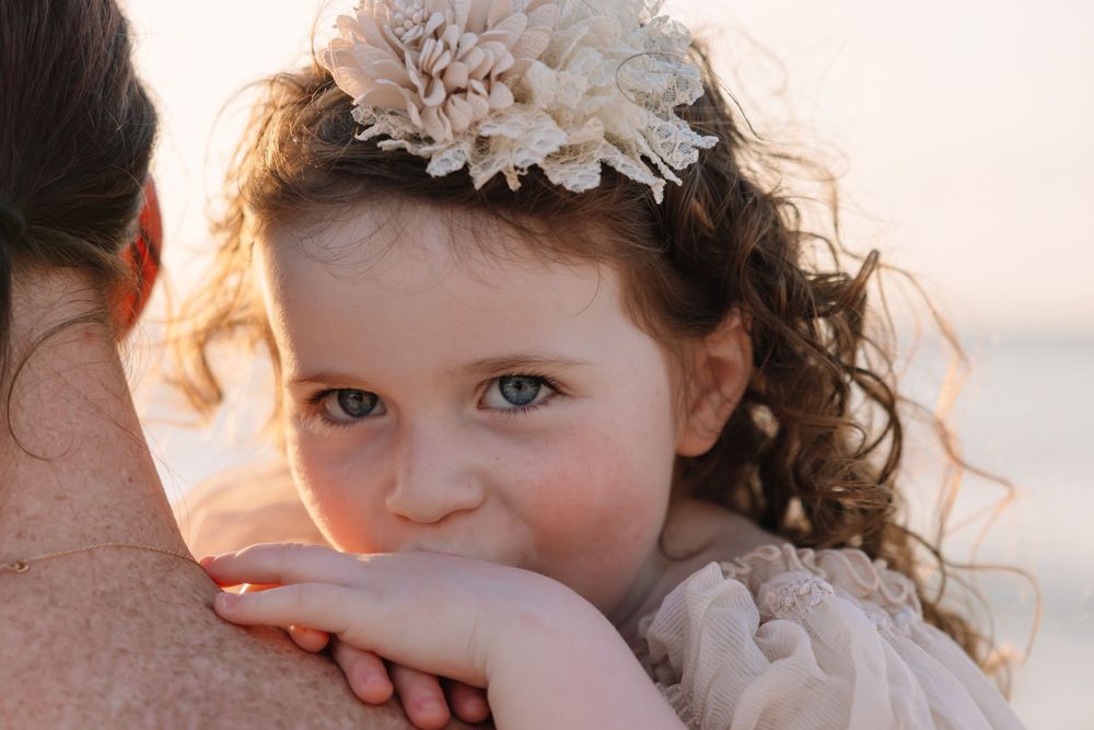 A young girl with curly hair and a floral headband rests her chin on an adult's shoulder at the beach.