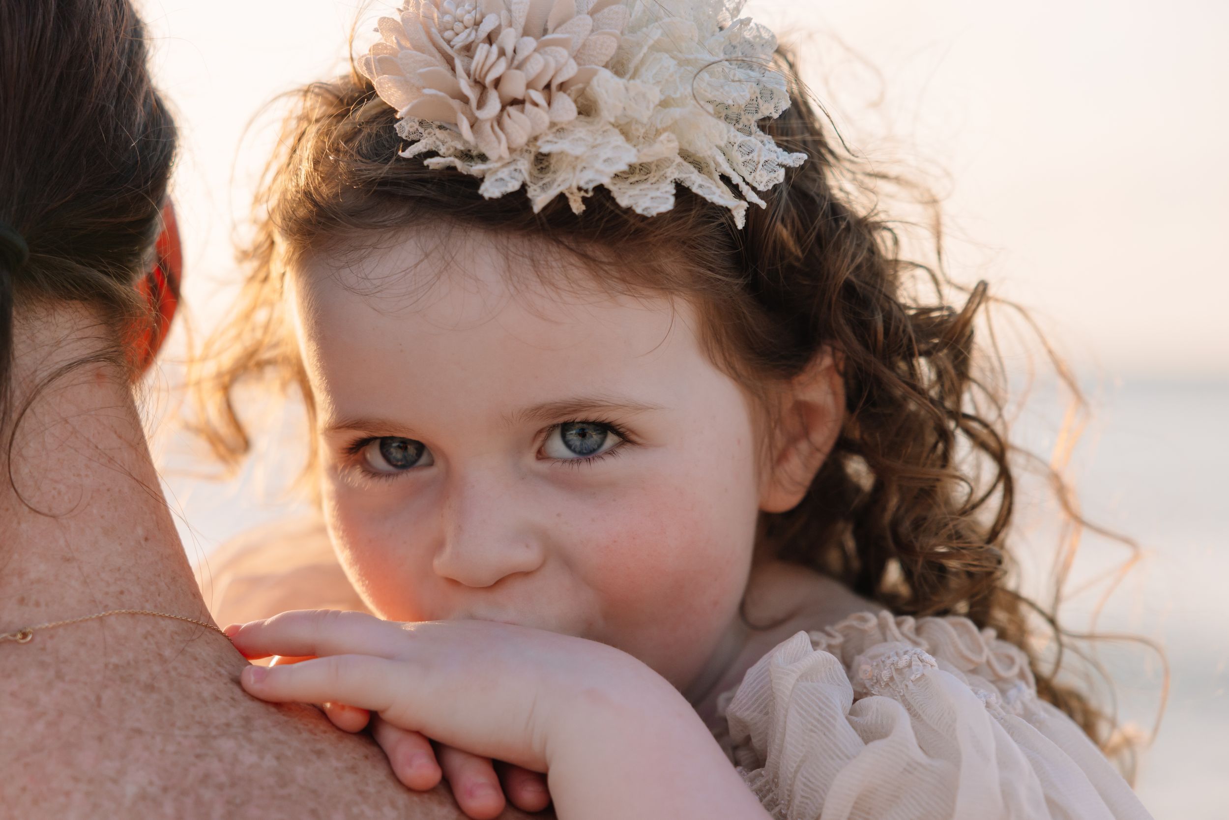 A young girl with curly hair and a floral headband rests her chin on an adult's shoulder at the beach.