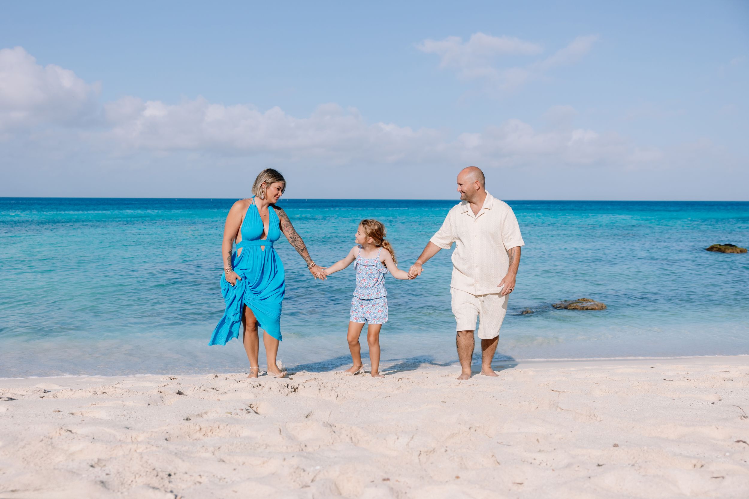 Family of three holding hands and walking along a white sand beach beside turquoise ocean water under a blue sky.