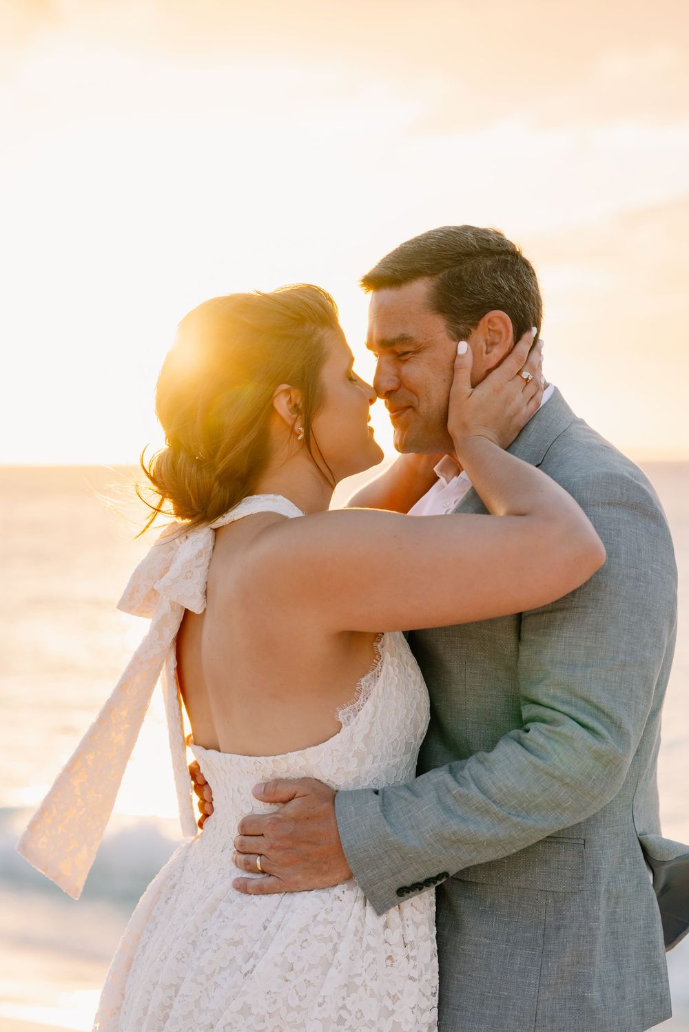Bride and groom share a romantic sunset kiss on the beach, she in a lace halter gown, he in a gray suit.