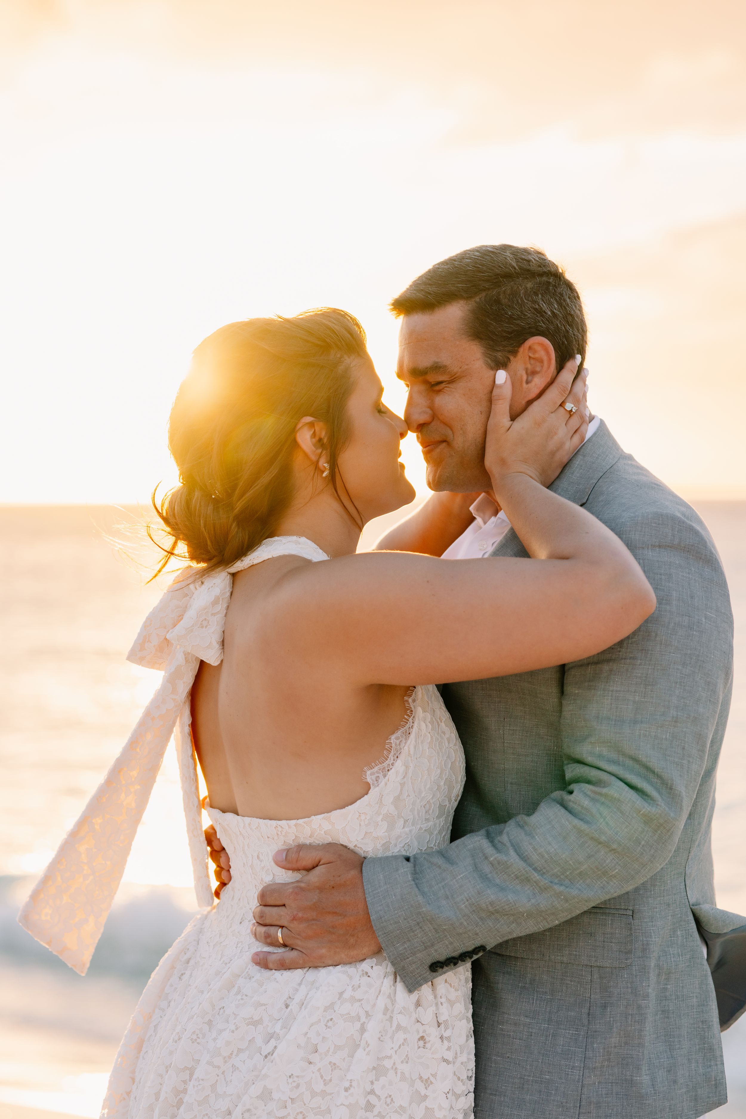 Bride and groom share a romantic sunset kiss on the beach, she in a lace halter gown, he in a gray suit.