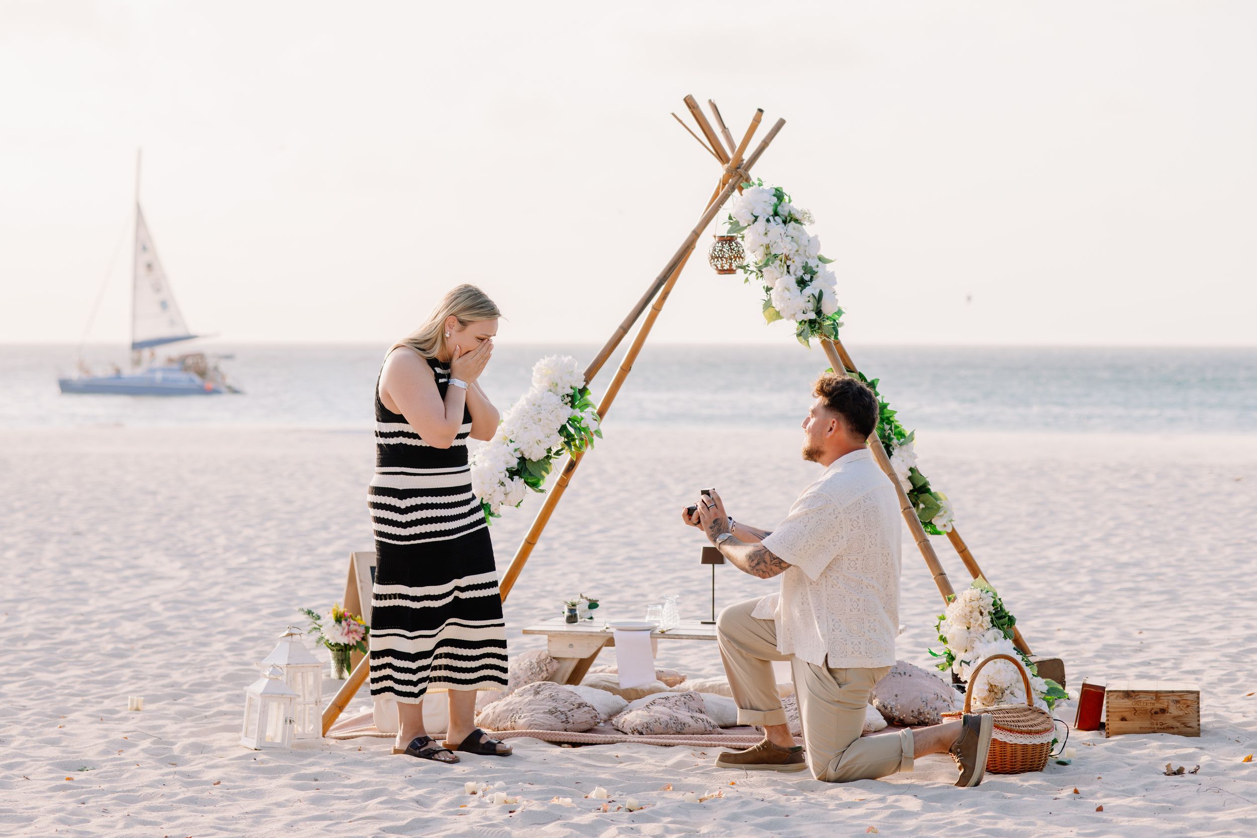 Man proposes to woman on beach with floral teepee backdrop, sailboat in distance, romantic picnic setup at sunset.