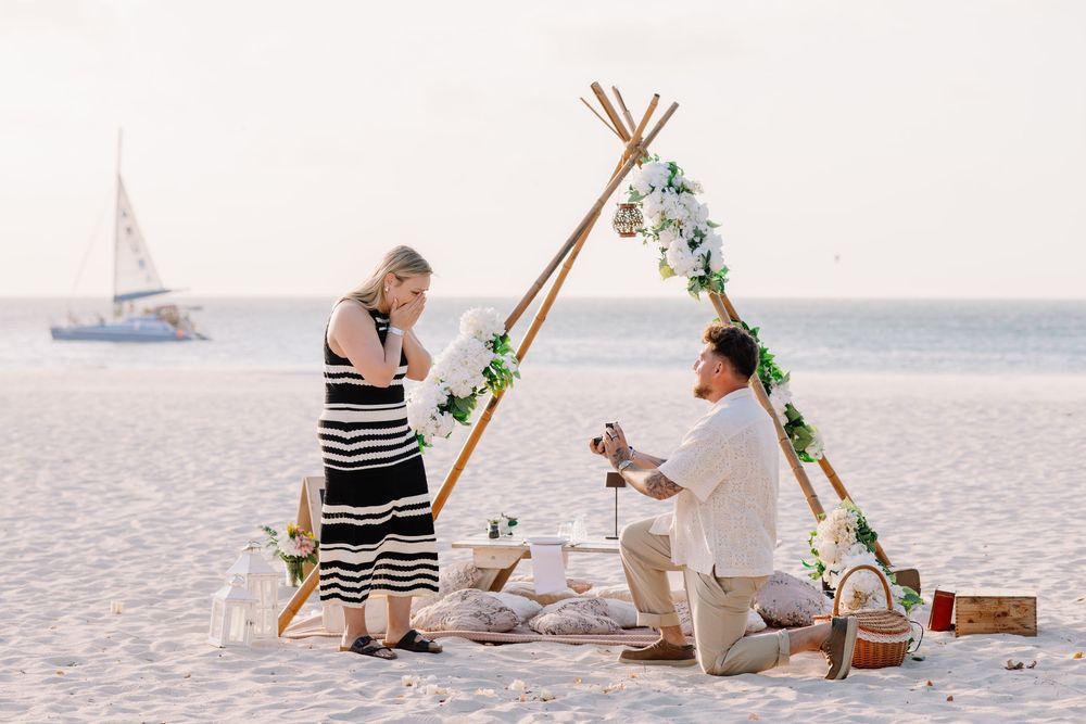 Man proposes to woman on beach with floral teepee backdrop, sailboat in distance, romantic picnic setup at sunset.