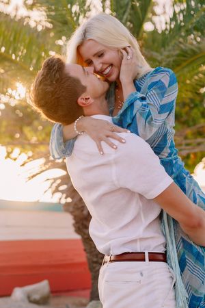 Couple sharing a romantic moment at sunset near a palm tree, man lifting woman in blue dress outdoors.