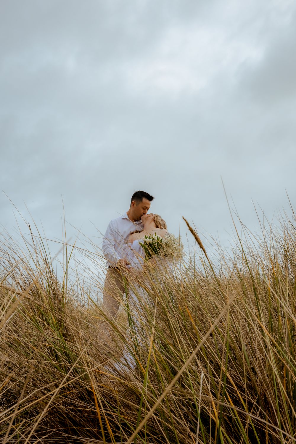 Couple walking through golden beach grass against cloudy sky.