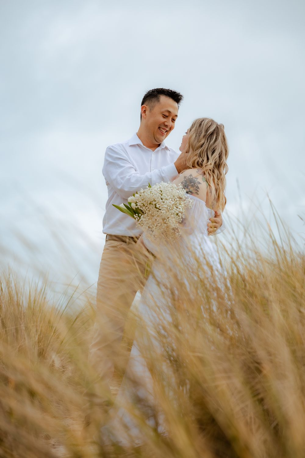 Couple in white and beige attire sharing intimate moment among beach grass.