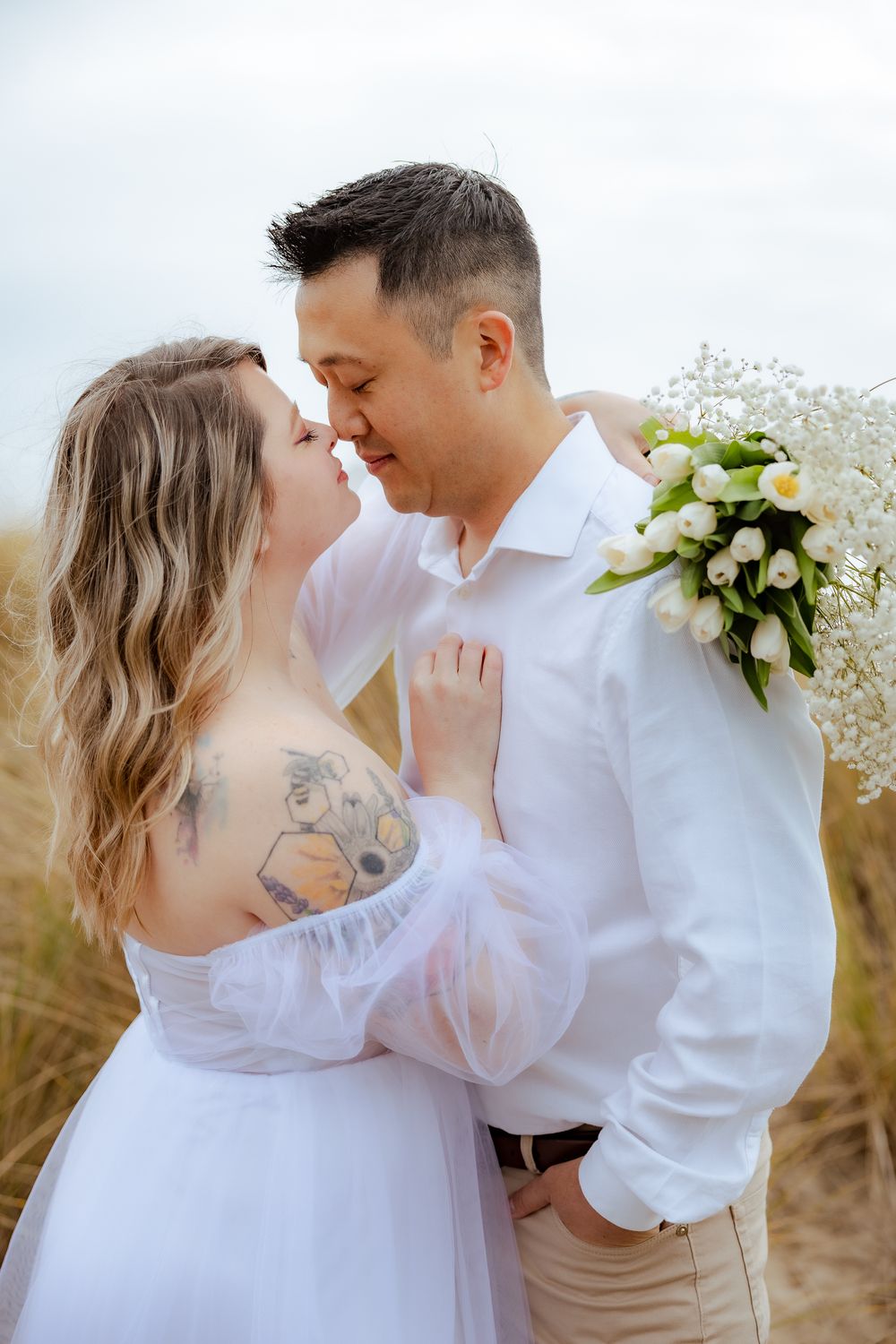 Couple in white clothing embracing with baby's breath bouquet against natural backdrop.
