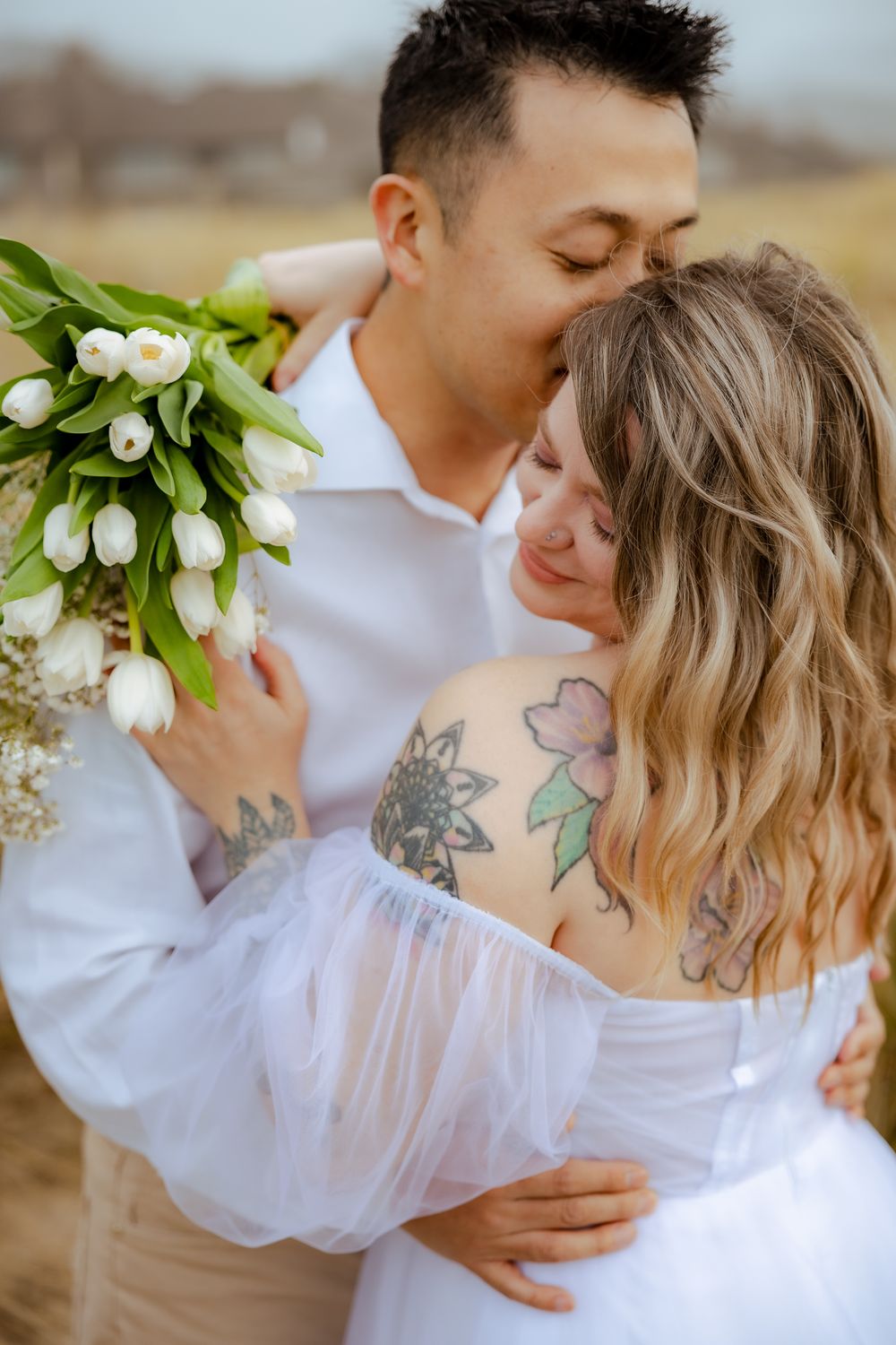 Romantic couple embraces in white outfits with white tulip bouquet against natural backdrop.