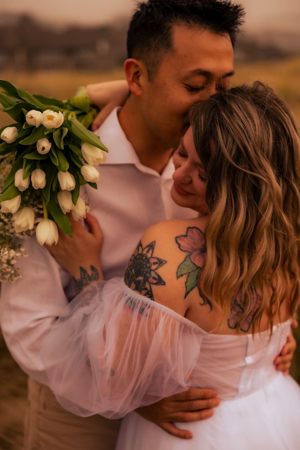 Romantic couple embraces while holding white tulips at sunset, with visible floral tattoos.