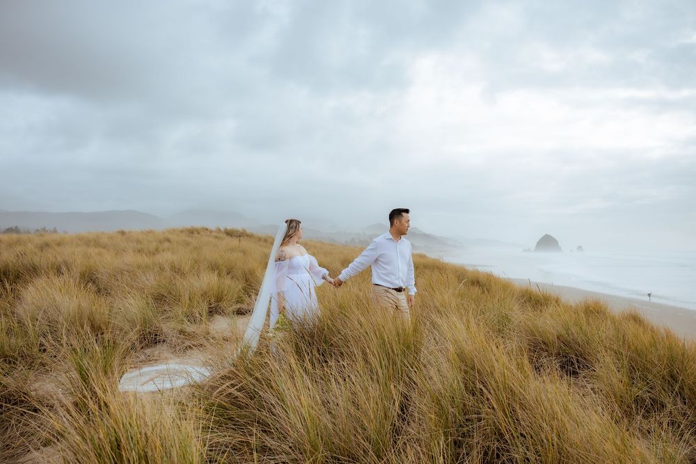 Couple walks through windswept coastal grass field under cloudy sky.