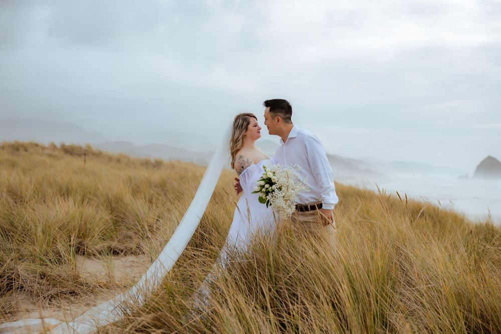 Couple shares tender moment in grassy coastal field with white bouquet.