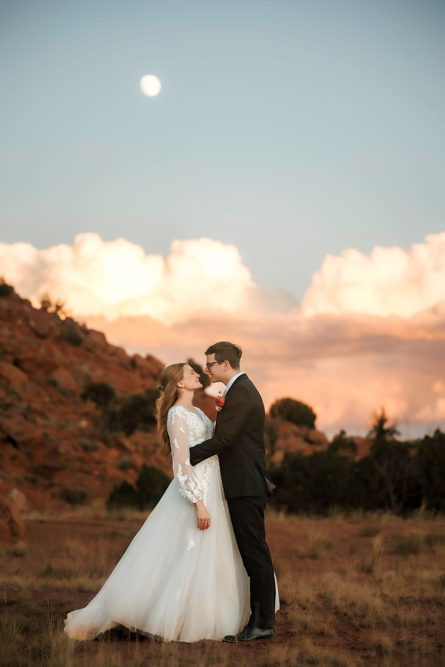 Bride and groom share a romantic moment under a full moon with dramatic sunset clouds behind them.