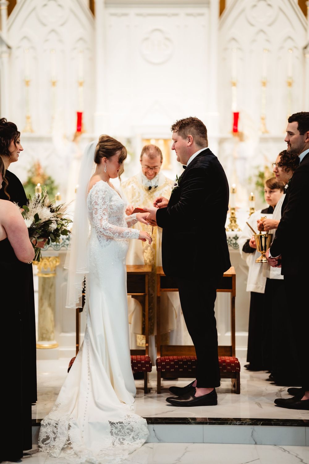 A wedding ceremony takes place in an elegant church with ornate white walls and glowing candles at the altar.