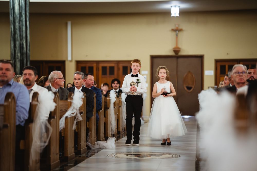 Young attendants walk down the church aisle during a wedding ceremony as guests look on from wooden pews.
