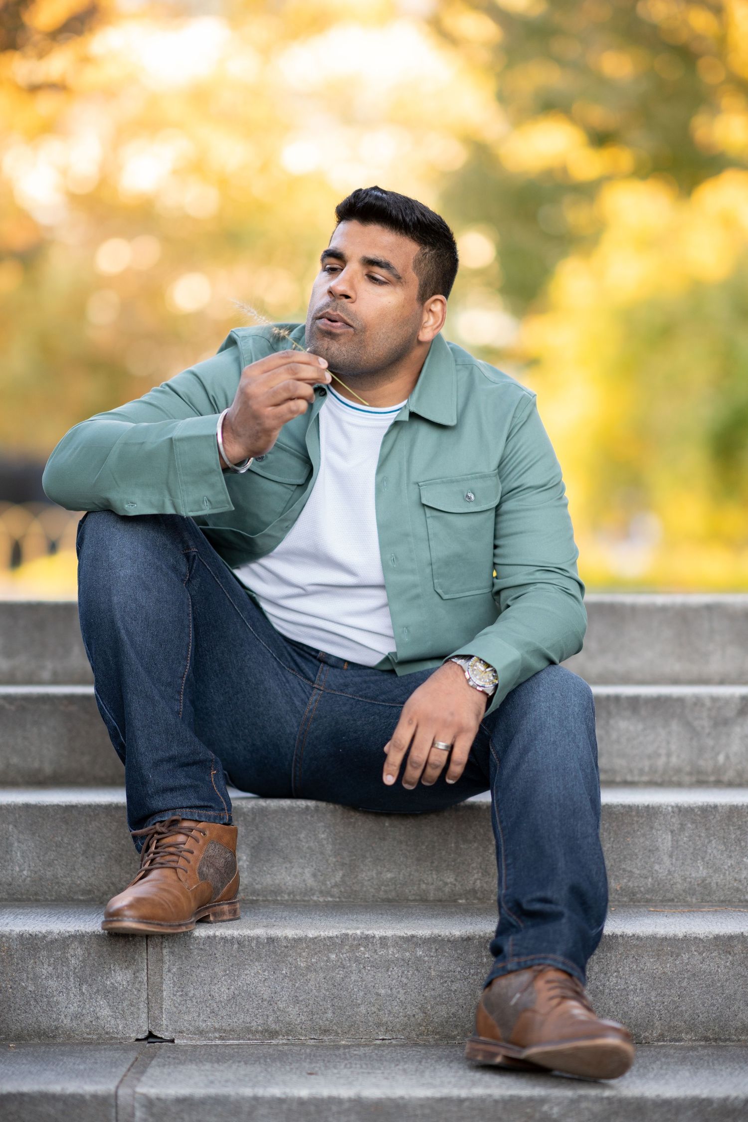 A person in a sage green jacket and jeans sits casually on concrete steps during golden hour.