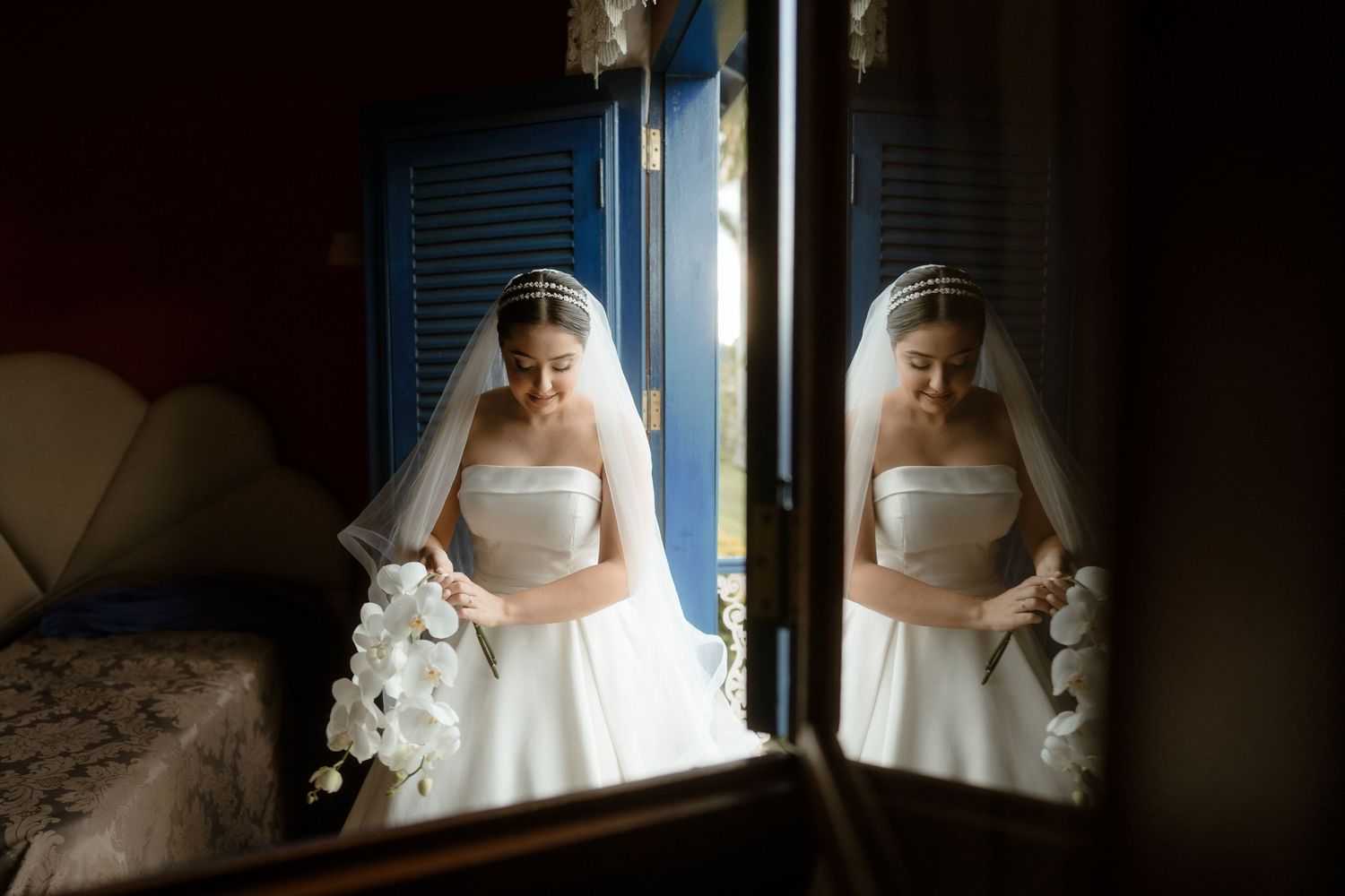 A bride in a white wedding dress stands by a window in dramatic lighting, creating a serene bridal portrait.