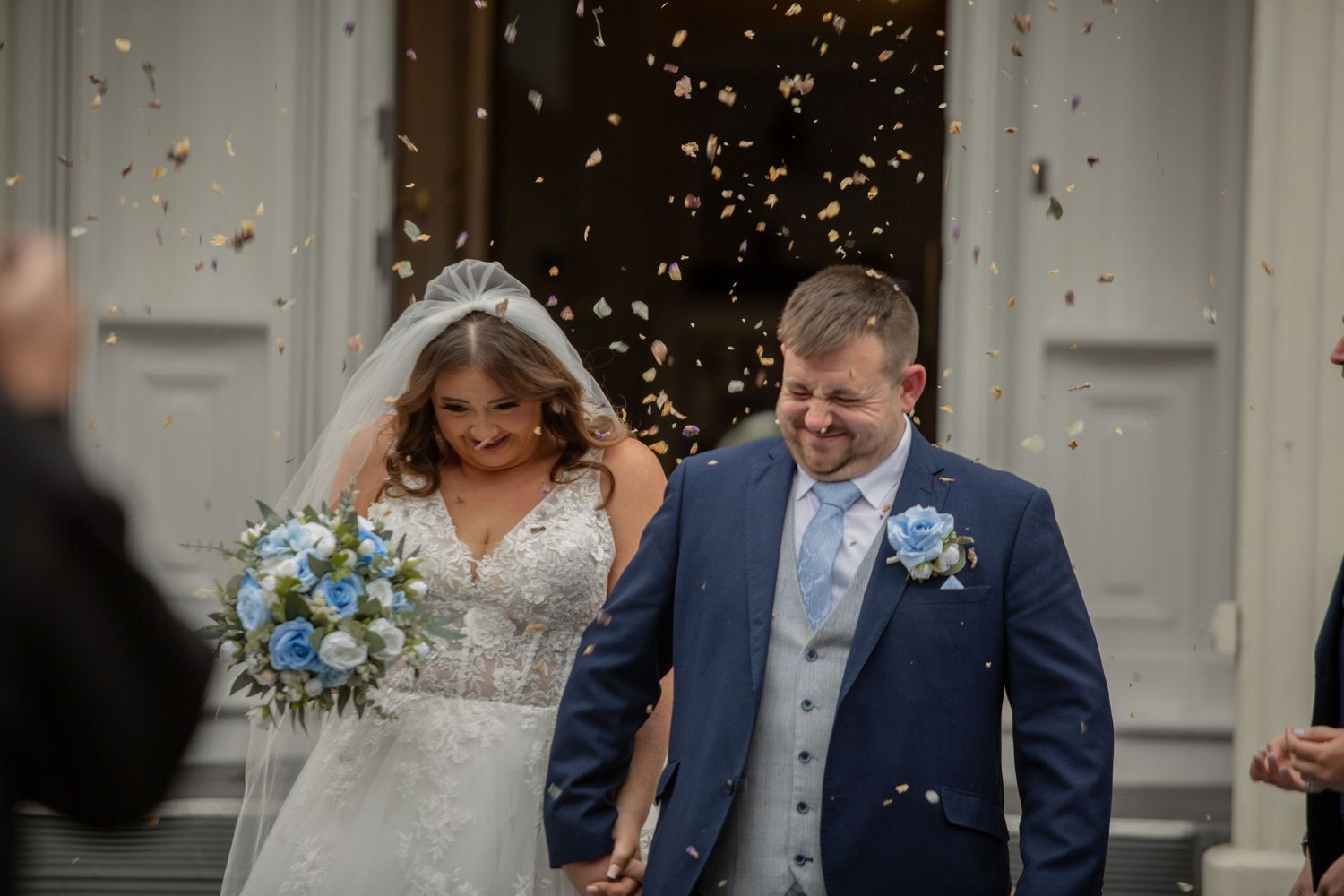 A joyful couple exits their wedding ceremony while guests throw confetti, with the bride holding a blue and white bouquet.