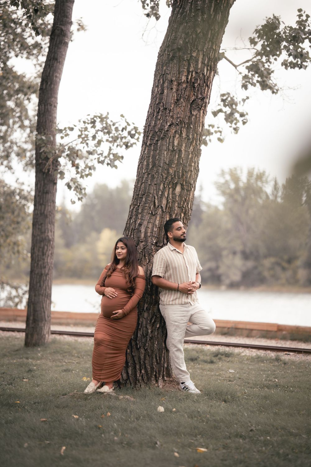 A couple in brown and white outfits pose playfully around a large tree trunk in a misty outdoor setting.