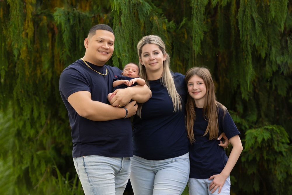 Family of four posing outdoors with baby against backdrop of weeping willow trees.