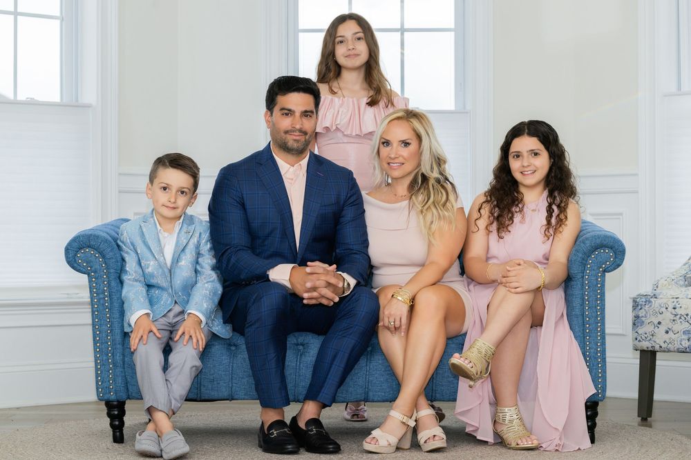 Family of five sitting together on blue couch in bright white room wearing coordinated pastel outfits.