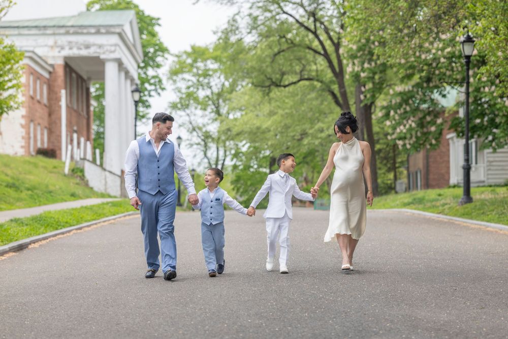Pregnant woman and man walk hand-in-hand with two young boys on a tree-lined path near a brick building.