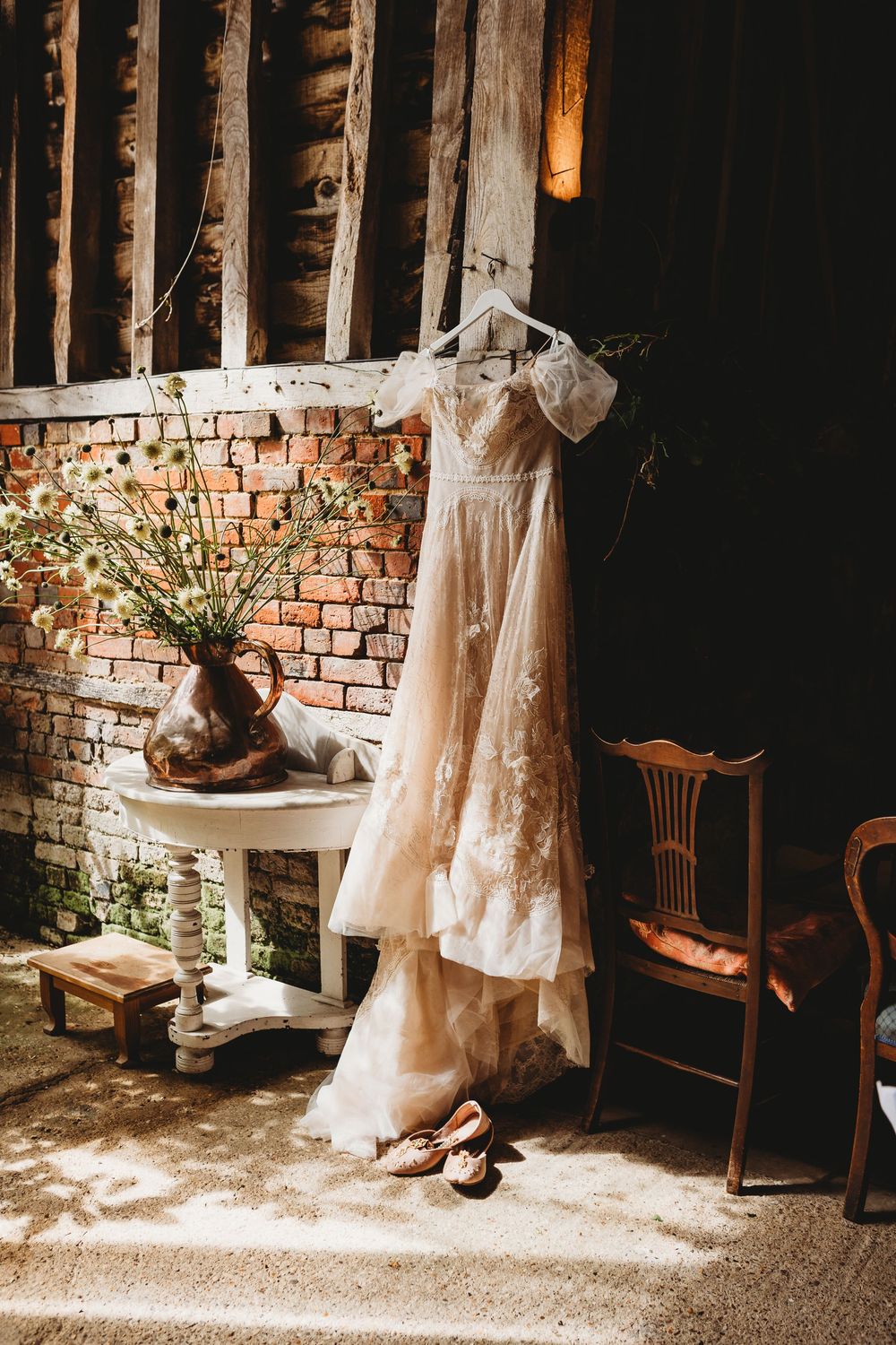 Vintage lace wedding dress hanging against exposed brick wall with floral arrangement and wooden chair.