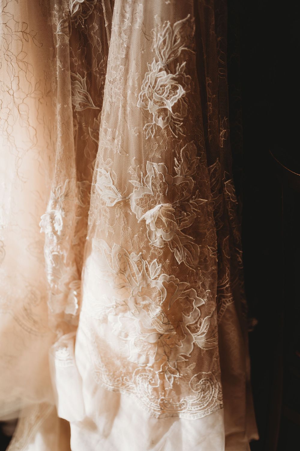 Close-up detail of intricate lace embroidery on a white wedding dress in warm sunlight.