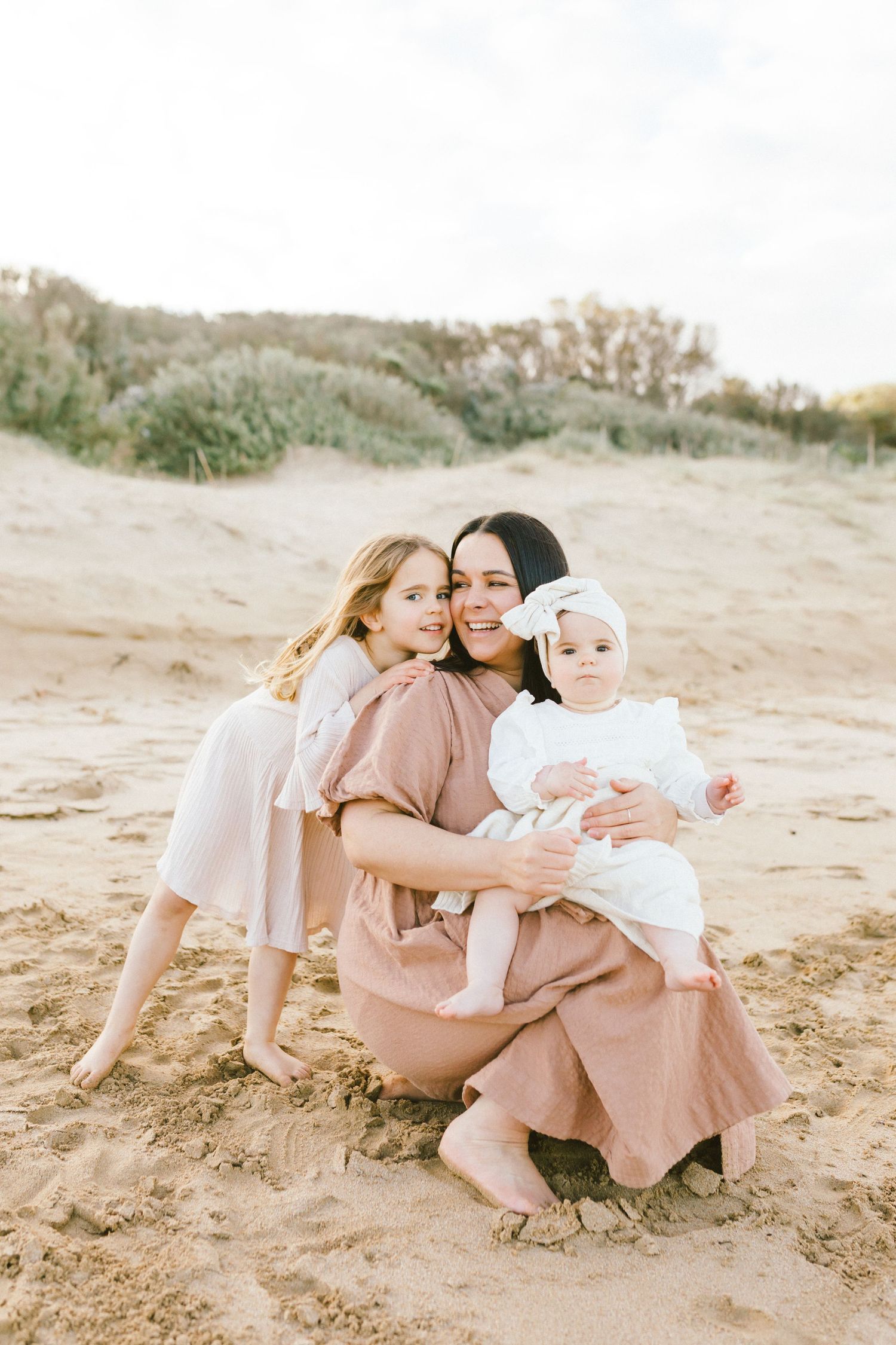 Family wearing matching blush pink dresses sits together on sandy beach during golden hour photo session.