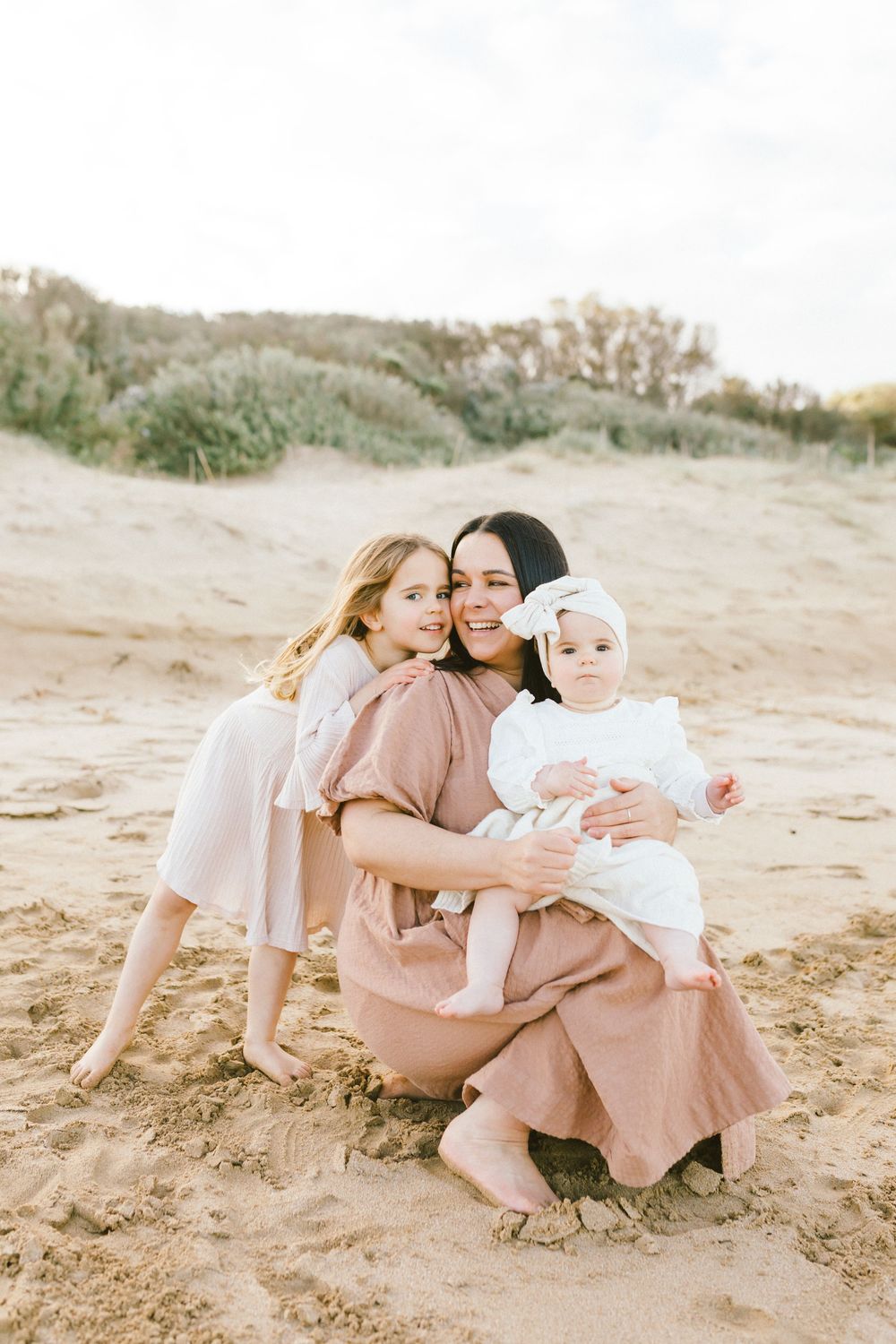 Family wearing matching blush pink dresses sits together on sandy beach during golden hour photo session.