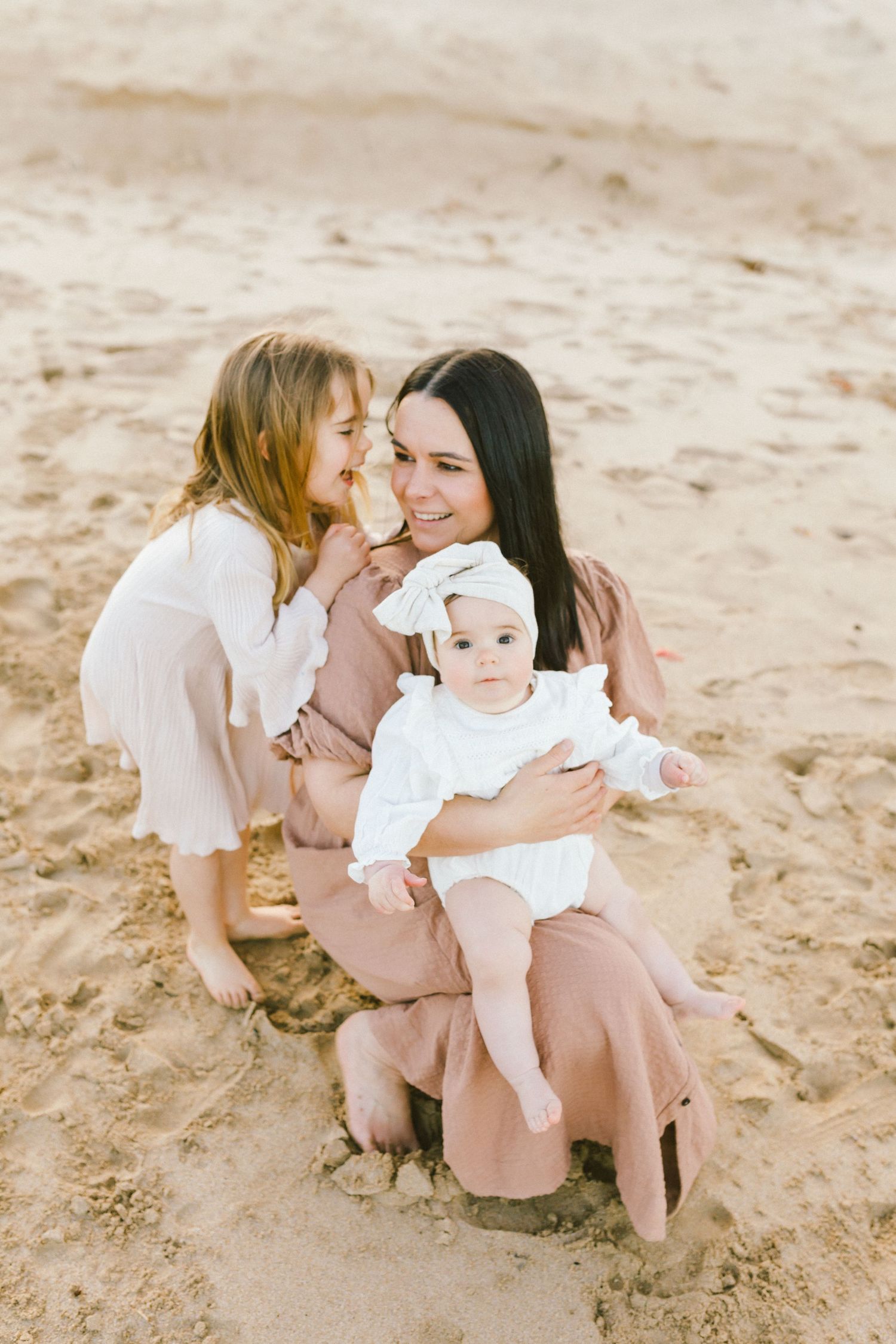 Family wearing white and beige outfits shares tender moments together on sandy beach in series of candid photos.