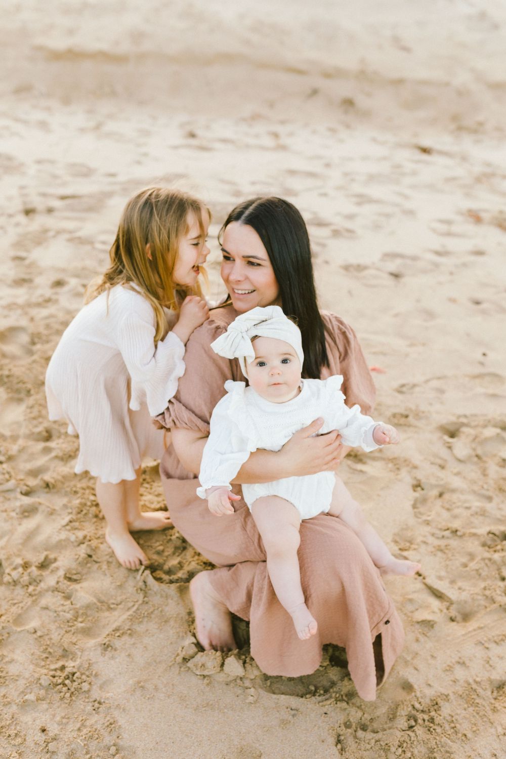 Family wearing white and beige outfits shares tender moments together on sandy beach in series of candid photos.