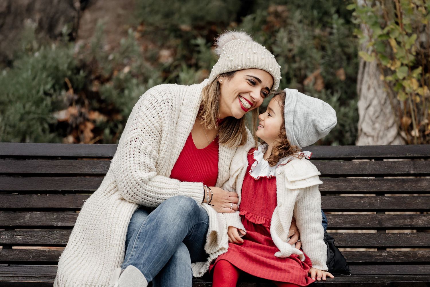 Two people wearing matching white sweaters and red tops share a sweet moment while sitting on a wooden bench outdoors.